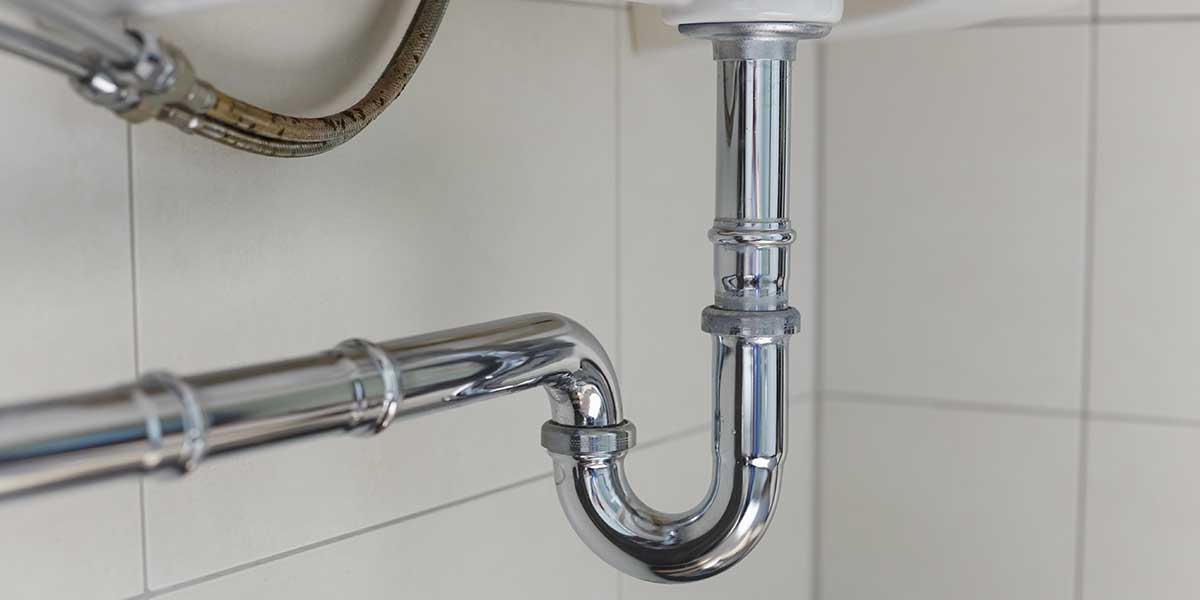 A chrome-plated metal P-trap pipe assembly installed underneath a bathroom sink against a white tiled wall.