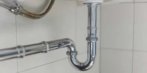 A polished chrome P-trap connects the drainage pipes beneath a bathroom sink against a white tiled wall.